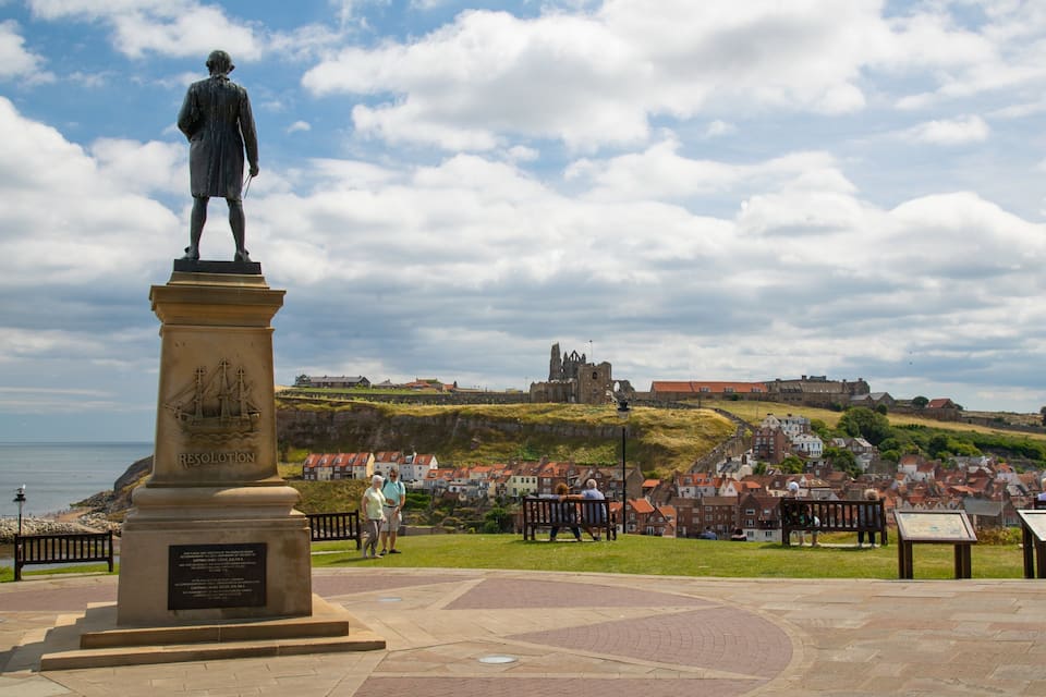 A statue of a historical figure on a pedestal with a plaque, overlooking a coastal town with a castle in the background.