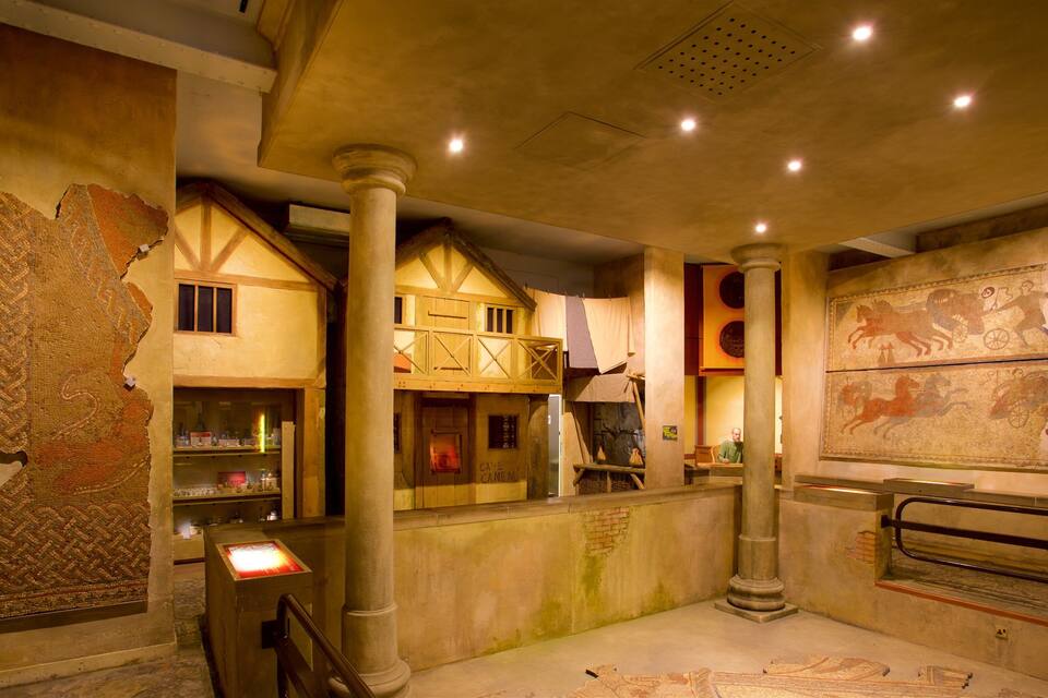 A museum interior with ancient artifacts, a model house, and a mural of a chariot race.
