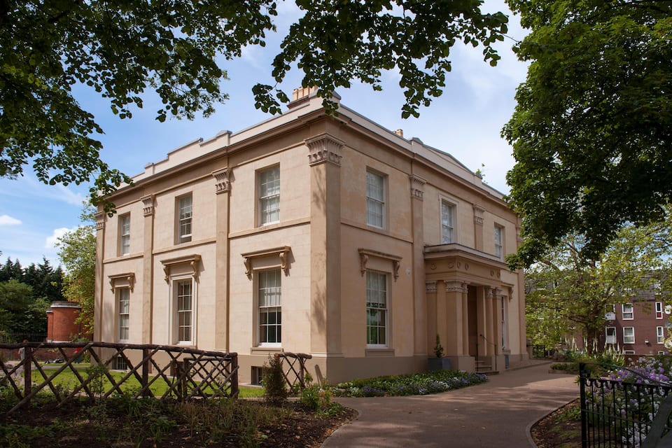 A historic building with classical architecture, a paved pathway, and surrounding greenery.