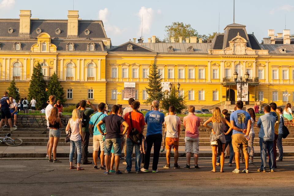 A group of people standing in front of a yellow building with classical architecture.