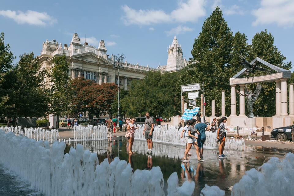 A fountain with water jets, a statue, and a historic building in the background.