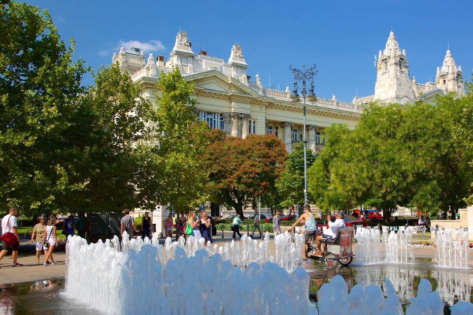Uma praça pública com uma fonte, árvores e um edifício grande e ornamentado ao fundo.