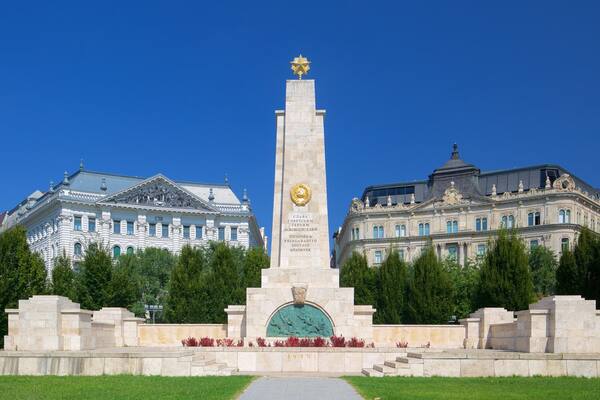 Un monument orné d’inscriptions, entouré d’arbres et avec deux grands bâtiments en arrière-plan.