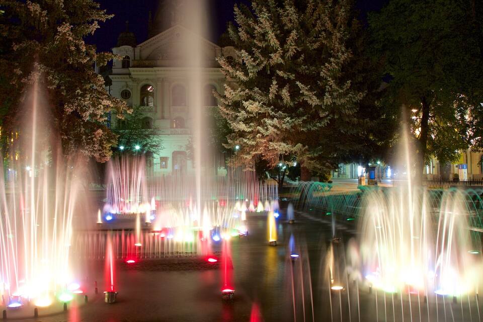 A nighttime fountain display with water jets illuminated by multicolored lights, set against a backdrop of a historic building and cherry blossom trees.