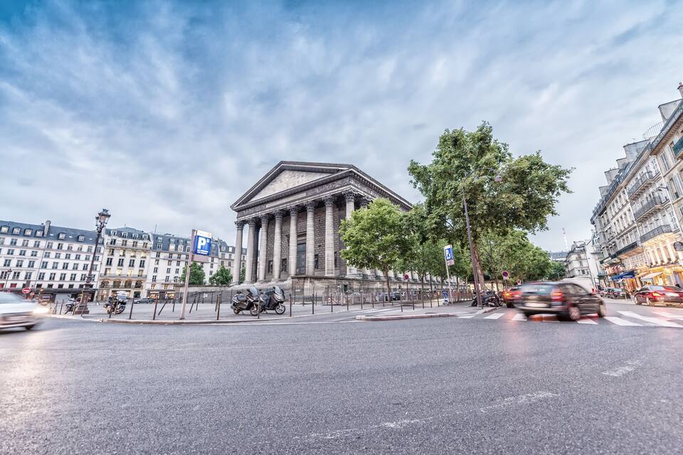 A busy intersection with a neoclassical building, parked scooters, and cars.