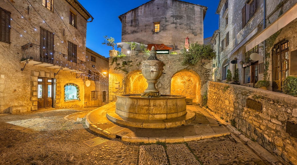 Ein historischer Innenhof aus Stein mit einer zentralen Fontäne, Kopfsteinpflasterwegen und traditionellen Gebäuden mit Rundbogenportalen.