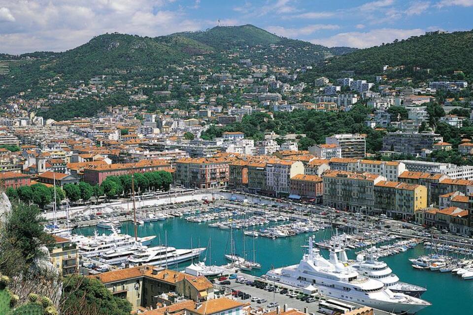 A coastal cityscape with a marina filled with boats, surrounded by terraced buildings and hills.
