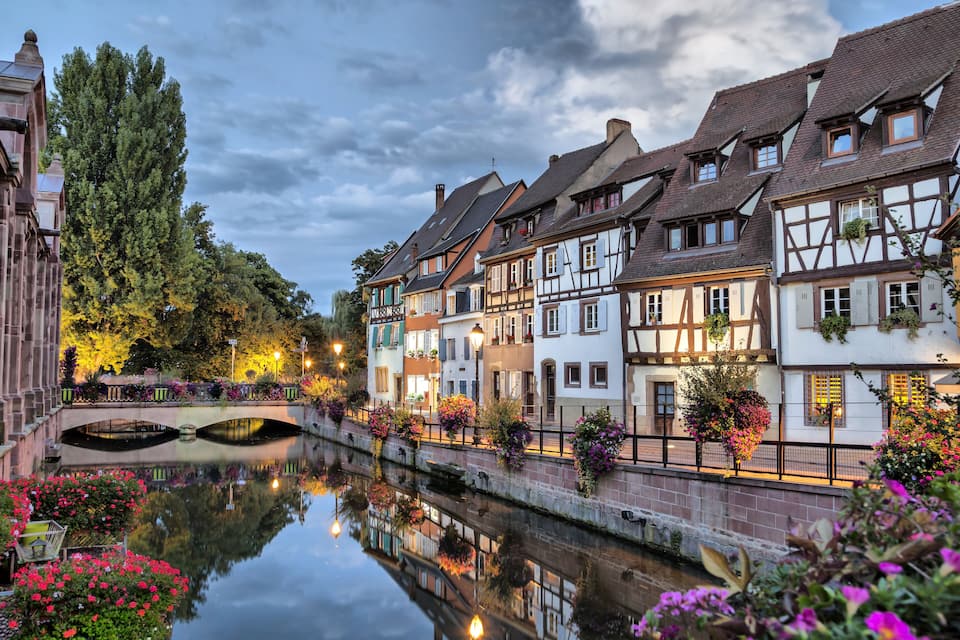 Ein Kanal mit sich im Wasser spiegelnden Häusern, umgeben von blühenden Pflanzen und Bäumen.