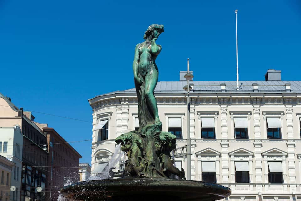 Une statue en bronze représentant une femme et un lion, placée dans une fontaine, avec un bâtiment de style classique en arrière-plan.