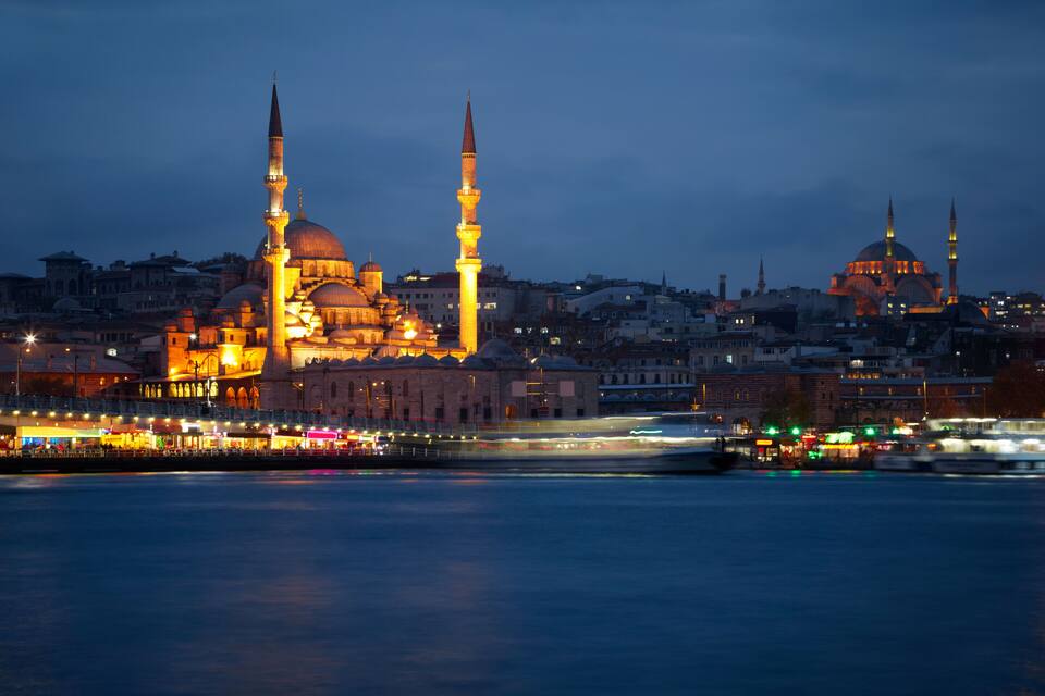 Paesaggio urbano al crepuscolo con cupole e minareti illuminati di una moschea, un fiume sullo sfondo e un ponte con luci.