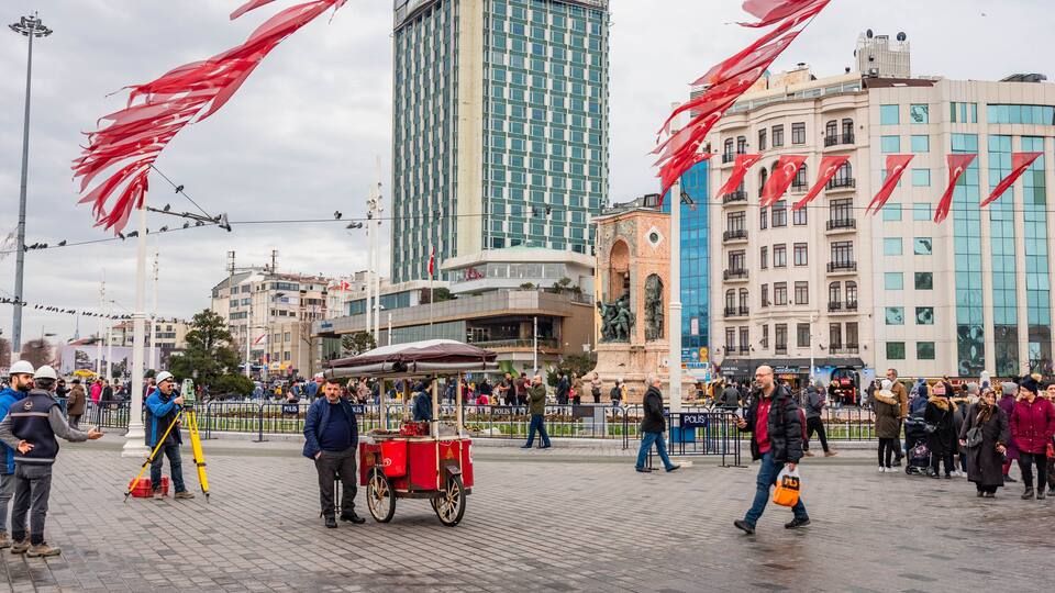 A busy urban square with a red kiosk, people walking, and modern buildings in the background.