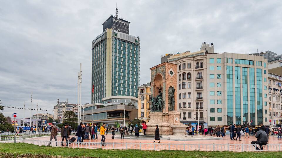 A busy urban square with a prominent clock tower, modern buildings, and pedestrians.