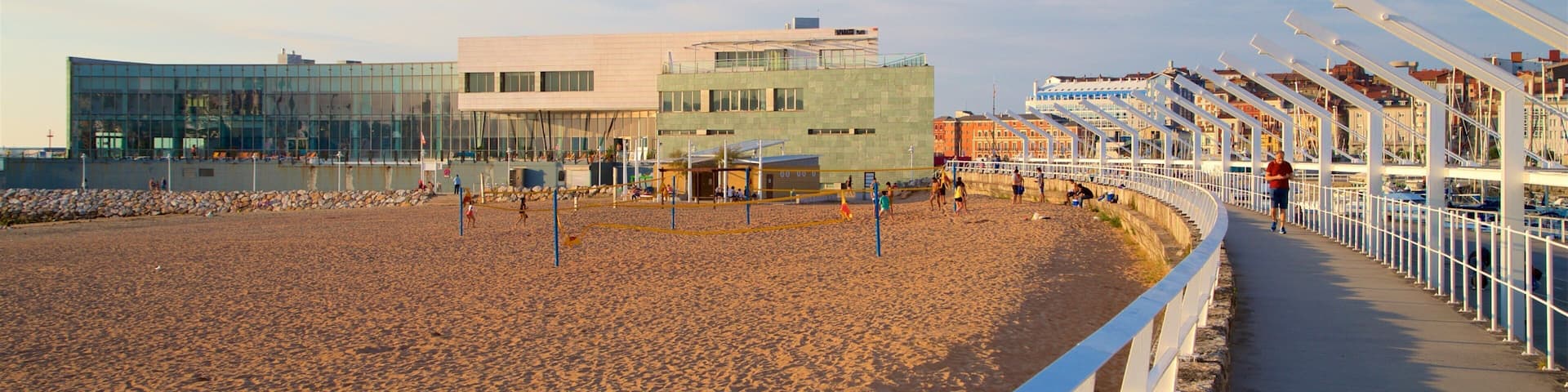 A sandy beach with a volleyball court, a modern building, and a white fence along the promenade.