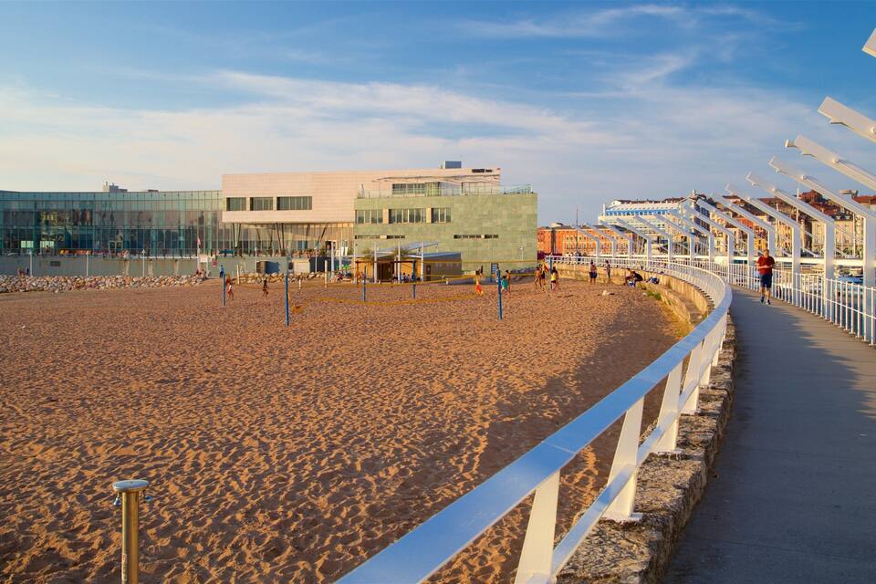 Una playa arenosa con una cancha de voleibol, un edificio moderno y una cerca blanca a lo largo del paseo marítimo.