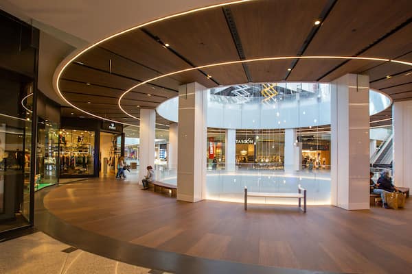 A modern shopping mall interior with wooden flooring, circular ceiling design, and glass storefronts.