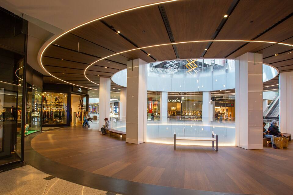 Interior de um shopping moderno com piso de madeira, design de teto circular e vitrines de vidro.