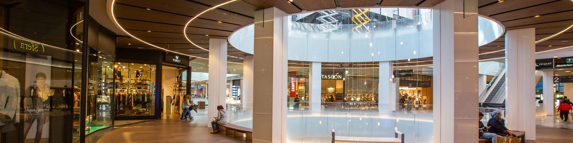 A modern shopping mall interior with wooden flooring, circular ceiling design, and glass storefronts.