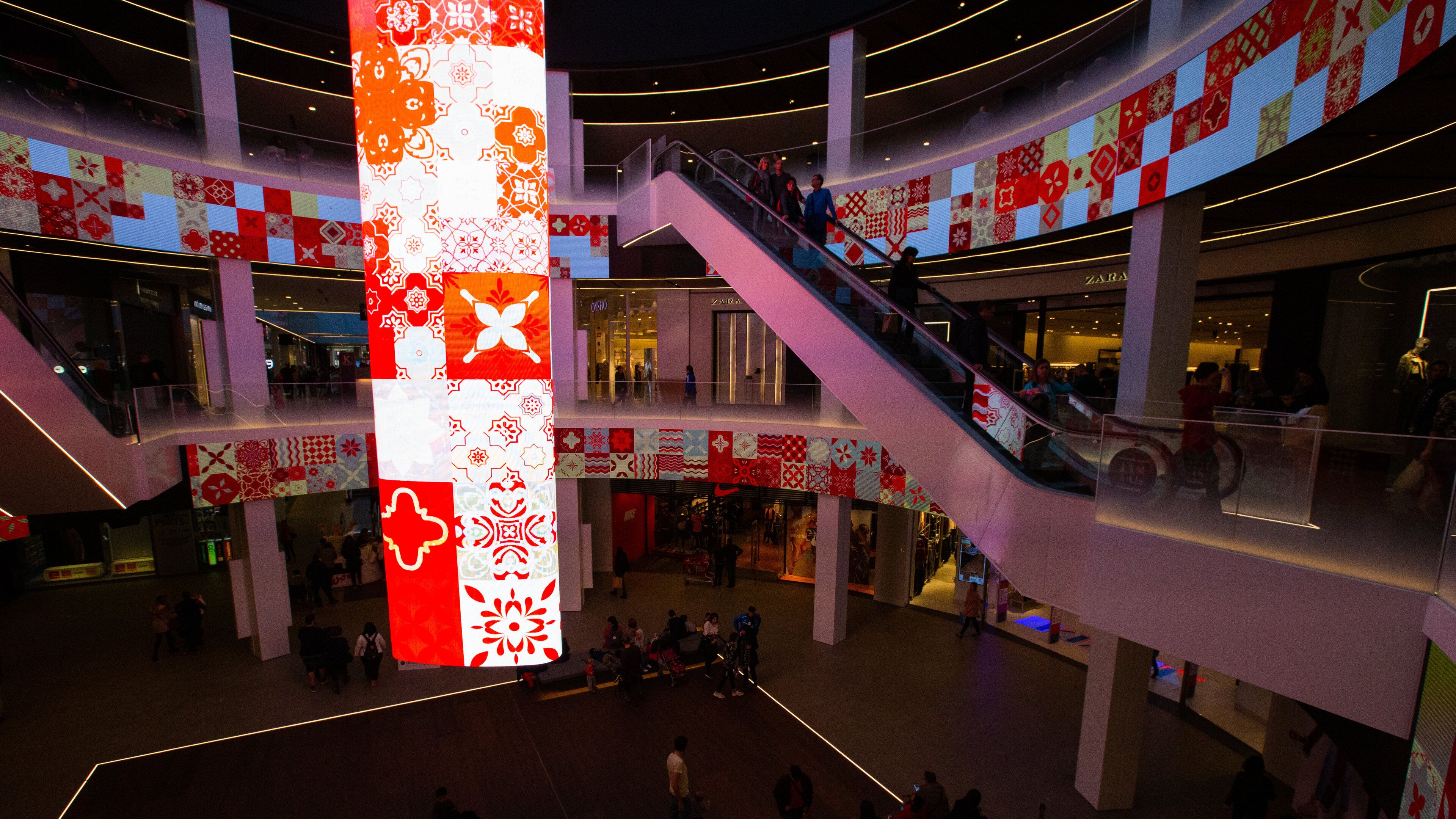Un centre commercial à plusieurs niveaux, doté d’escalators, d’un grand écran vertical illuminé et de divers magasins, dont Zara.
