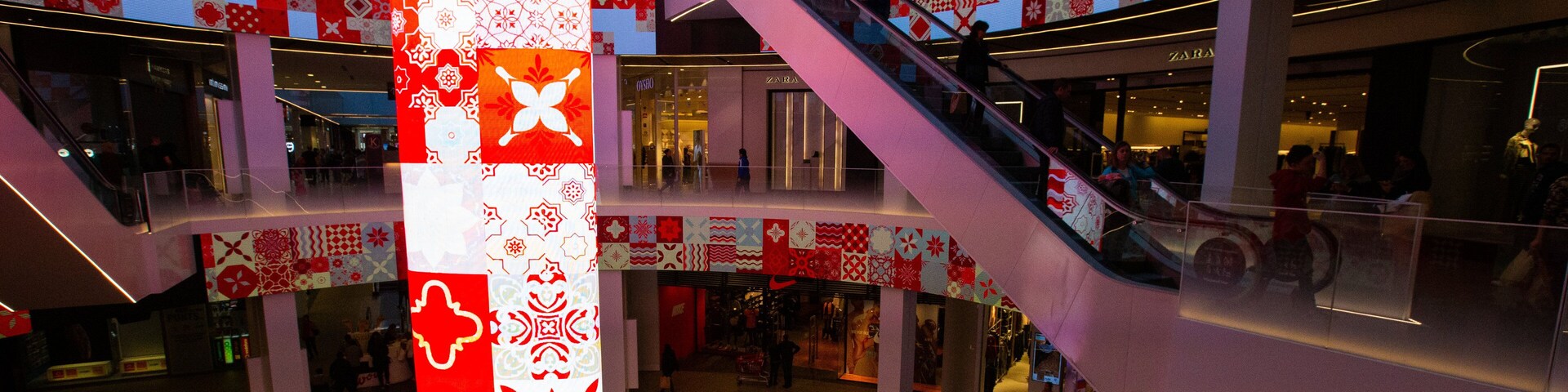 Um grande centro comercial com vários níveis, escadas rolantes, um painel vertical iluminado e diversas lojas, incluindo a Zara.