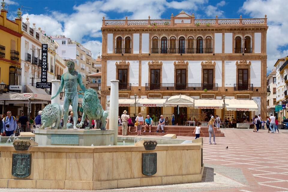 A plaza with a statue of a man and two lions, surrounded by historic buildings and cafes.