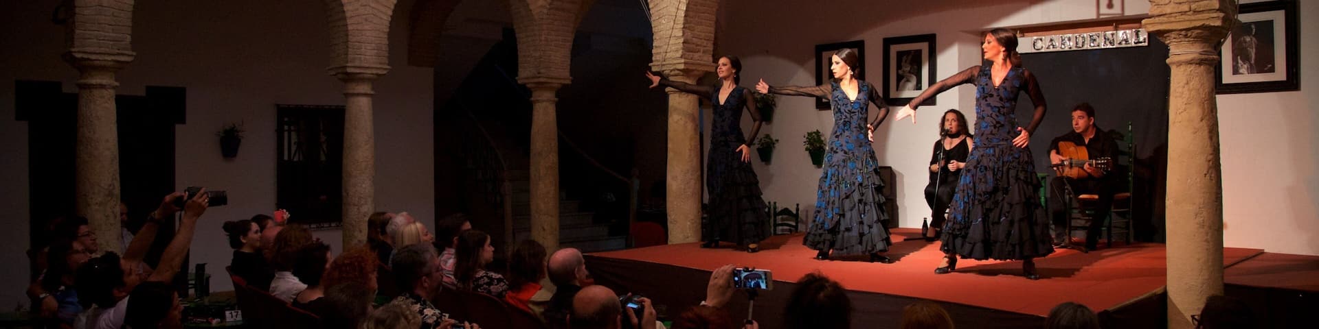 Un escenario con artistas vestidas de oscuro y un hombre tocando la guitarra, rodeado por un público en un patio interior con arcos.