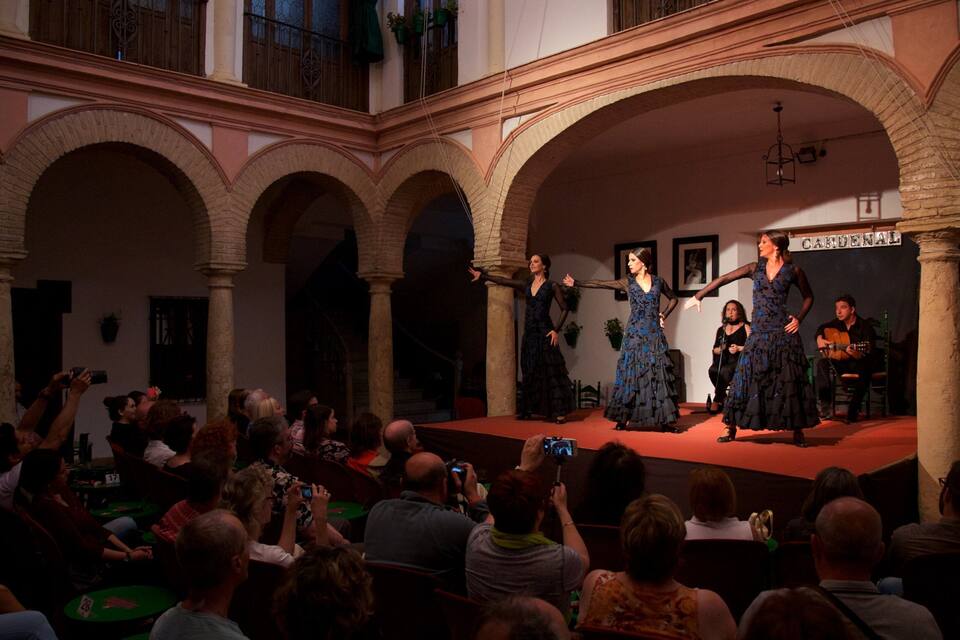 A stage with performers in dark dresses and a man playing guitar, surrounded by an audience in an indoor courtyard with arches.