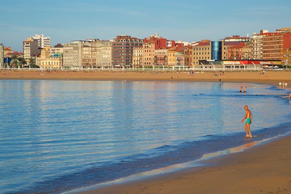 Een strand met helderblauw water, een zandstrand en een stadsgezicht met kleurrijke gebouwen op de achtergrond.
