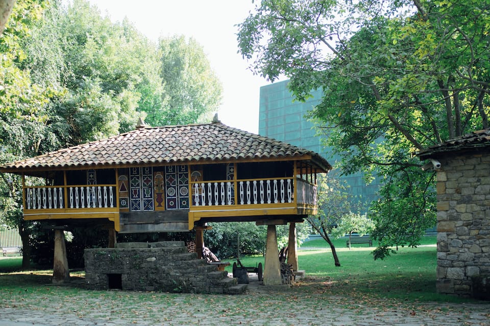 En traditionell paviljong av trä med ett tak täckt av tegelpannor, omgiven av frodig grönska och en mur av sten.