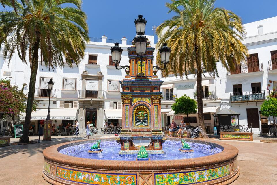 A plaza with a fountain, palm trees, and white buildings with balconies.
