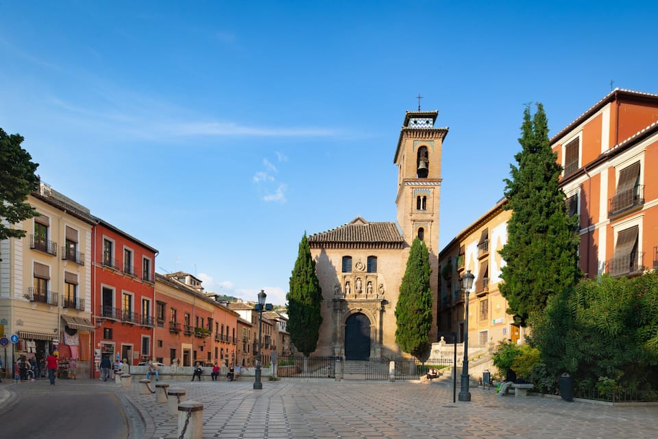 A cobblestone square with historic buildings, a prominent church tower, and a statue in the center.