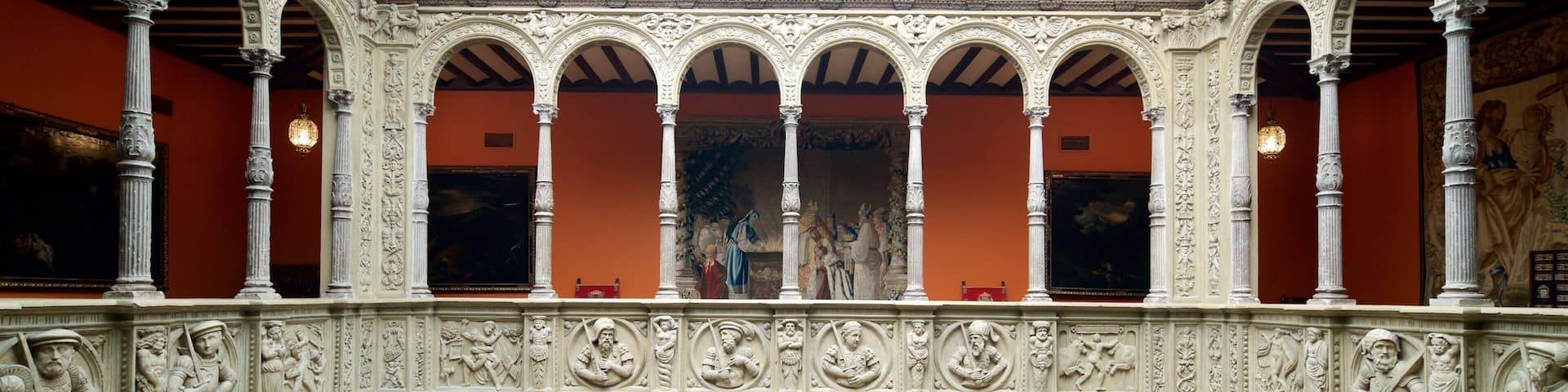 An ornate gallery with white columns and arches, featuring classical sculptures and framed artworks on the walls.