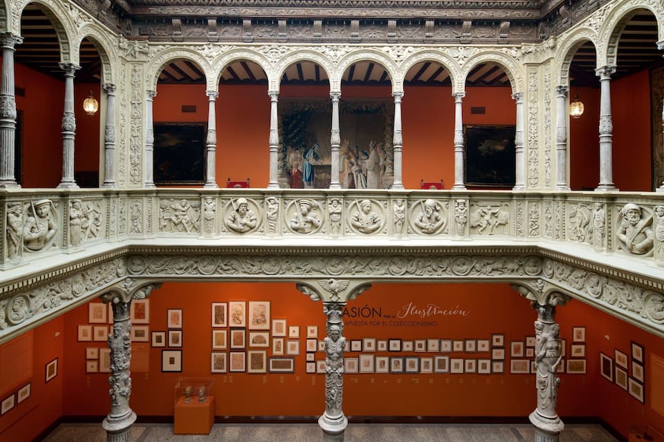 Uma galeria ornamentada com colunas e arcos brancos, exibindo esculturas clássicas e obras de arte emolduradas nas paredes.