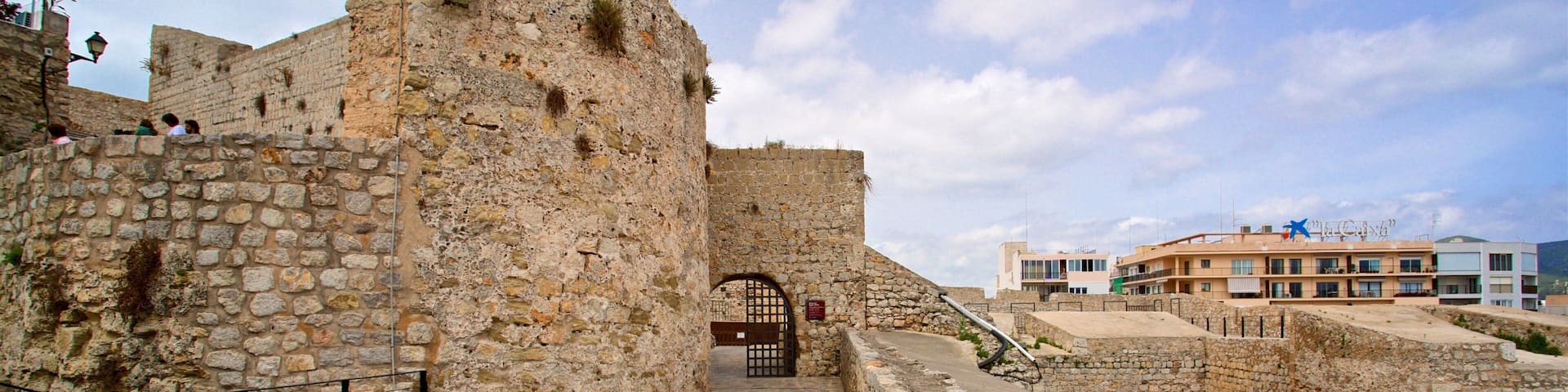 Una fortaleza de piedra con una puerta, un camino adoquinado y edificios modernos al fondo.