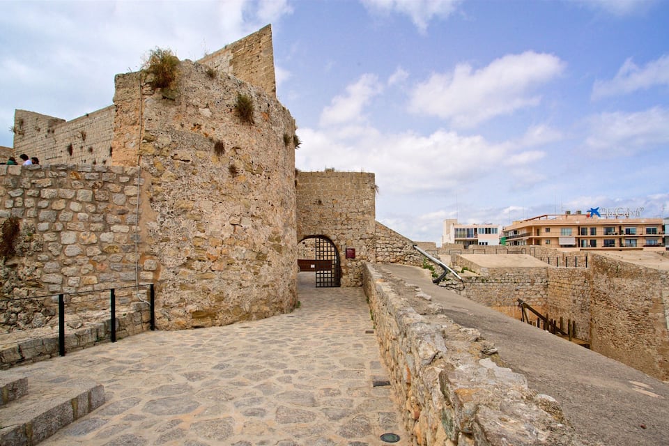 A stone fortress with a gate, a cobblestone path, and modern buildings in the background.