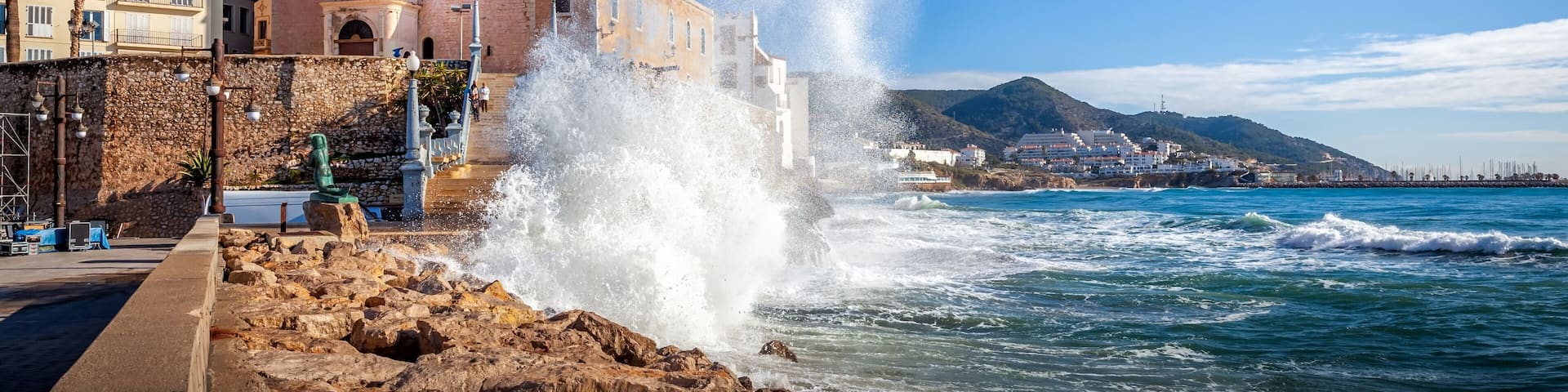 Una iglesia costera con un campanario, olas rompiendo contra las rocas y un cielo azul despejado.