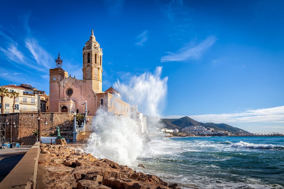 A coastal church with a bell tower, waves crashing against the rocks, and a clear blue sky.