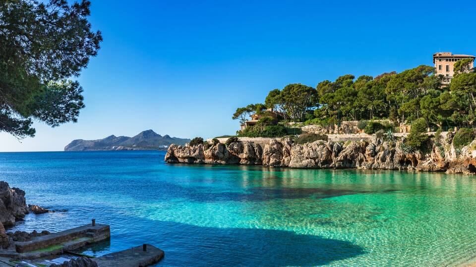 A coastal landscape with clear turquoise waters, a sandy beach, rocky cliffs, and a white building with a stone entrance.