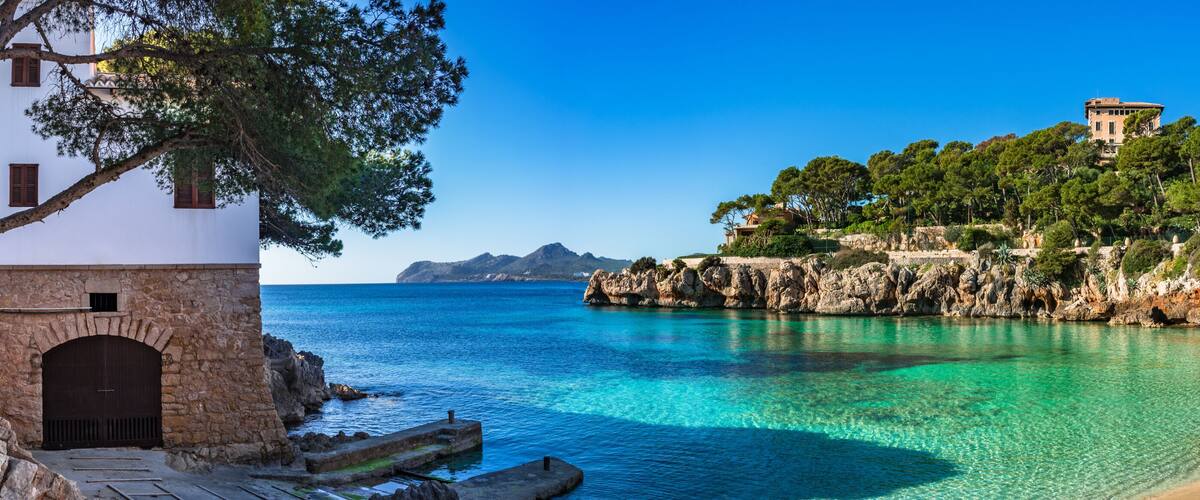 A coastal landscape with clear turquoise waters, a sandy beach, rocky cliffs, and a white building with a stone entrance.