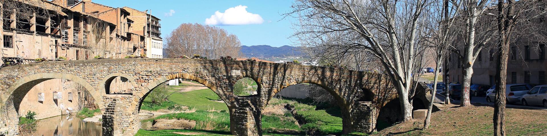 A stone bridge over a calm river, surrounded by grassy areas and trees.