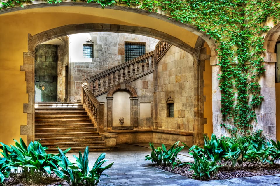 A stone staircase with a wooden railing leading up to a door, surrounded by greenery and arches.
