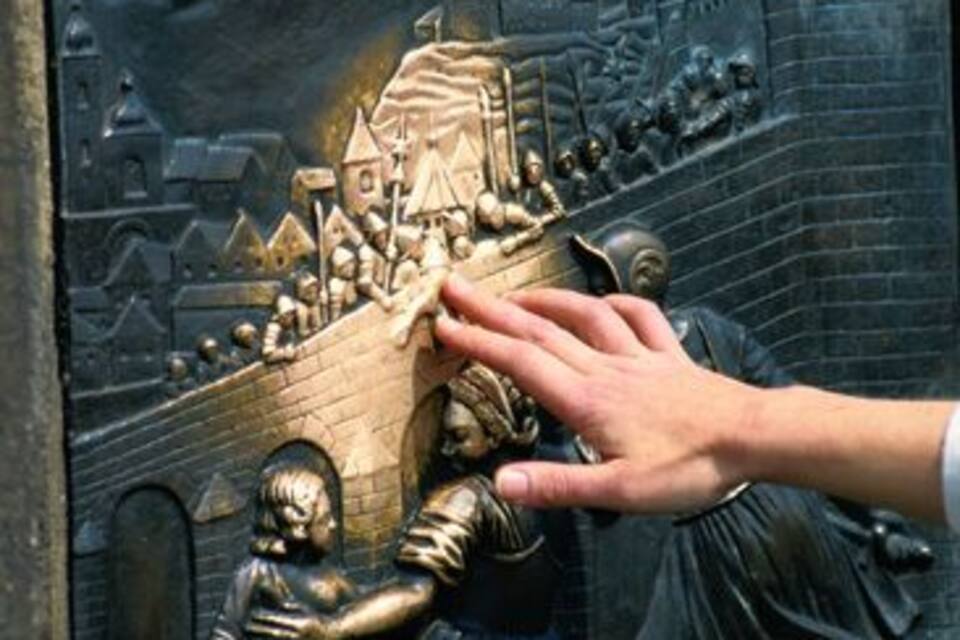A bronze relief sculpture with a hand reaching towards a figure in a robe, set against a backdrop of a cityscape with buildings and a bridge.