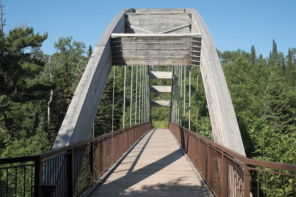 A wooden pedestrian bridge with metal railings, surrounded by dense forest.