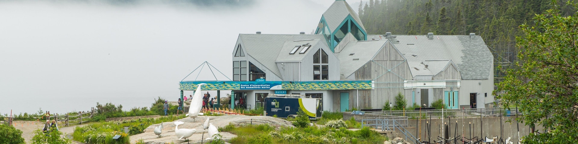 A coastal building with a pointed roof, surrounded by greenery and parked cars.