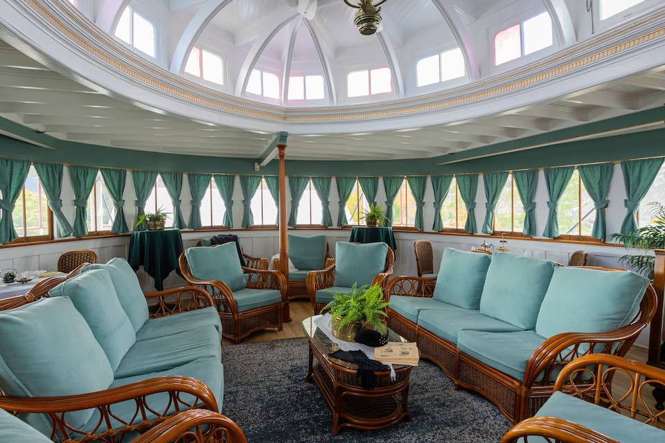 A room with a high, arched ceiling and large windows with green curtains. There are wicker chairs and a sofa with light blue cushions.
