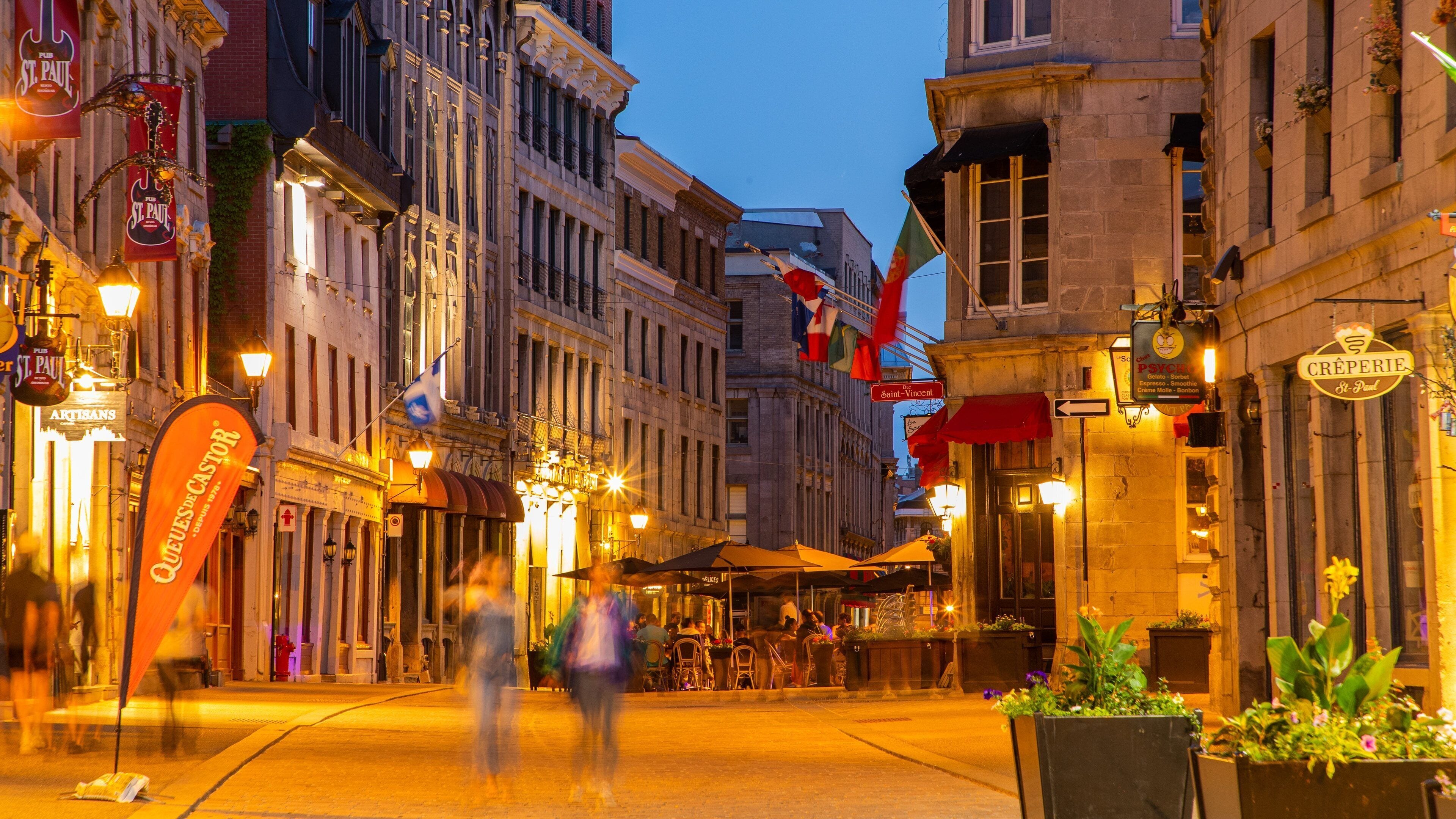 Eine belebte Straße im Dämmerlicht mit beleuchteten Schildern und Wimpeln, darunter 'St. Paul' und 'Creperie St. Paul'.