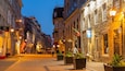 Una calle empedrada bordeada de edificios históricos, mesas al aire libre y plantas en macetas.