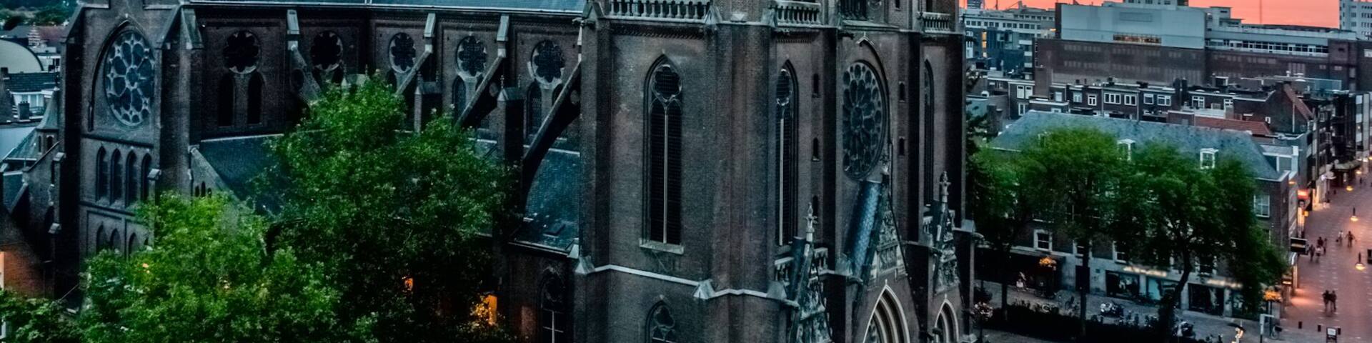 Une cathédrale gothique, dotée de deux flèches jumelles et d’une tour horloge, entourée d’immeubles et d’arbres.