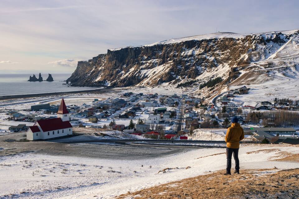 A snowy coastal town with a church, houses, and a person standing on a hill overlooking the sea.