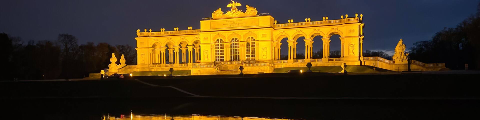 A grand illuminated building with a reflection in the water, featuring a central archway and statues.
