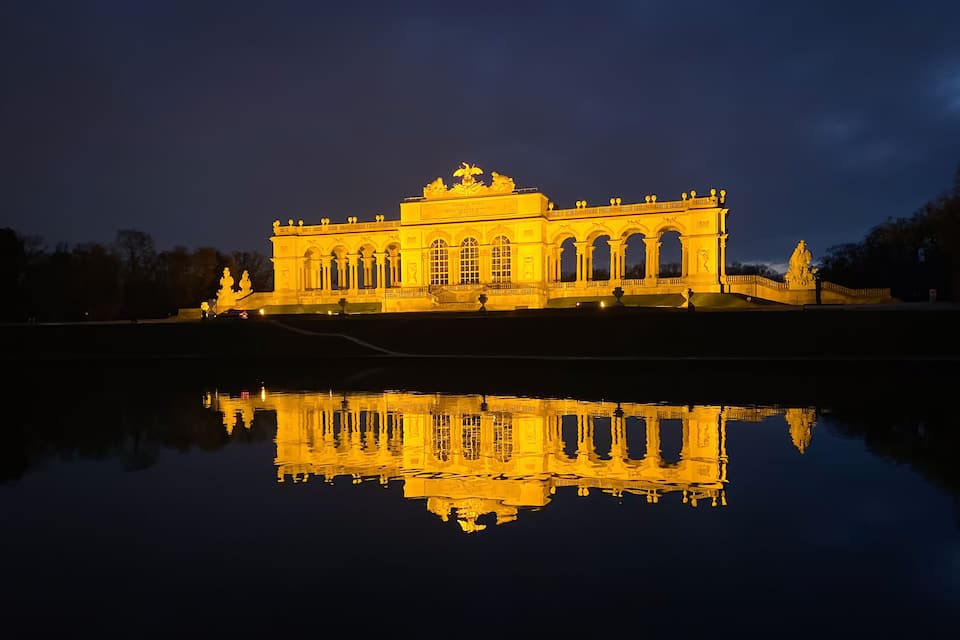 Un edifici majestuós i il·luminat amb el seu reflex en l'aigua, que destaca un arc central i estàtues.